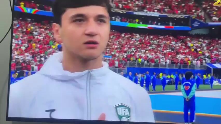 This media file captures a moment on a screen, likely a television, showing a sporting event. In the foreground, the heads and shoulders of three individuals are visible. The person on the left is a young man with dark hair, wearing a white hooded jacket with a green and silver crest that reads "UFA". He is looking slightly off-camera with a neutral expression. To his right is another man, also wearing a similar white hooded jacket. He has dark hair and is looking to the right with his mouth slightly open, as if speaking. The third person, on the far right, has lighter hair and a beard, and is looking to the right with a thoughtful expression.

Behind these individuals, a large stadium crowd is visible, dressed predominantly in red and white. They appear to be spectators at a match. In the middle ground, a line of people in bright blue uniforms are standing on a blue surface, possibly a field or a stage. One person in a teal uniform with a heart emblem on their back stands out. The atmosphere appears to be during daylight hours, and there are no obvious indicators of weather conditions.

Visible text includes a logo in the top right corner that says "ZOR TV" with "LIVE UZB" below it. There are also signs in the background that appear to say "FIFA WORLD CUP". The overall scene suggests a pre-game or post-game interview or presentation at a major football event. Given the location context of Mashrab, Uzbekistan, this likely depicts a sporting event with Uzbek participation or broadcast. The emotions displayed by the foreground subjects are varied, ranging from neutral to engaged, suggesting they are either involved in the event or being interviewed.
