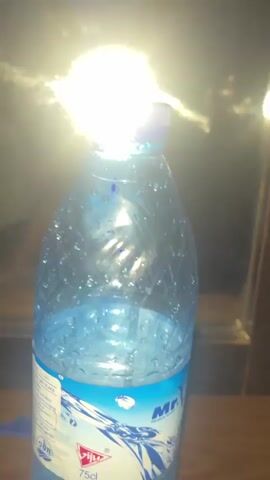 A video captures a blue-capped plastic water bottle with water droplets on its surface, illuminated by a bright light source causing strong lens flare and reflections. The camera pans out and around the bottle, revealing a second similar water bottle partially on the right and a grey sleeve or arm on the left, all within a dark, indistinct environment.