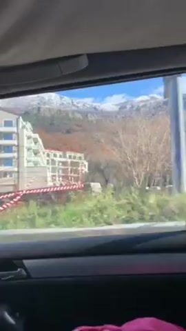 The video shows a view from inside a moving vehicle. The camera is positioned looking out the side window. The vehicle is traveling over a blue railing bridge. Beyond the bridge is a landscape with trees and greenery. A building with multiple balconies is visible on the left, and in the distance, a mountain range with snow-covered peaks is seen. The sky is blue. The video is shaky due to the vehicle's motion.