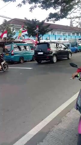 A street scene outside a blue building with flags. There are several vehicles including cars and motorcycles. People are visible walking and riding motorcycles. The overall impression is of a busy street in front of an institution, possibly a school, with Indonesian flags indicating a national holiday or celebration.
