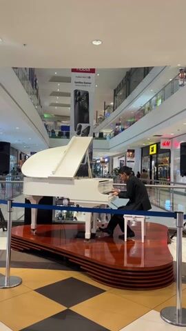 A man in a black suit sits at a white grand piano, playing it in the middle of a shopping mall. The piano is on a multi-tiered wooden platform, and there are two black speakers on stands flanking the piano. Blue rope is set up around the platform to create a barrier.  The mall is multi-storied, with shops visible in the background. An advertisement for a Nubia phone hangs above the piano. Mall patrons are seen walking around on the various levels of the mall. The floor is shiny with a tile pattern.