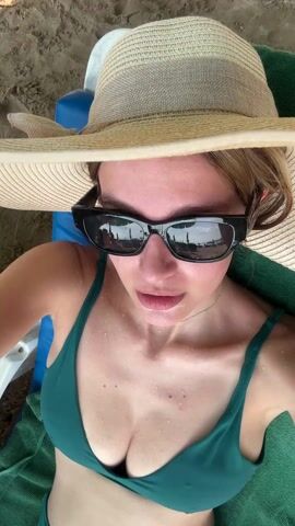 A woman wearing a green bikini, sunglasses, and a straw hat is lying on a beach chair. She adjusts her hat. The sunglasses reflect the beach scene with beach umbrellas. She smiles slightly. The camera is close to her face.