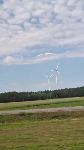 The video shows a scenic view of a rural landscape. In the foreground, there is a grassy area with some wildflowers and a fence line. A road is visible running horizontally across the middle of the frame. In the background, there are fields of crops, a line of trees, and several large wind turbines with their blades turning. The sky is blue with scattered white clouds. The camera appears to be moving, likely from a vehicle, as the landscape is captured from a side-view perspective.