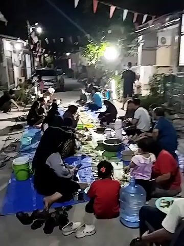 A group of people are gathered outdoors at night, sitting on blue tarps spread on the ground. They appear to be sharing a meal, with plates and food items visible. Some are eating, while others are talking or observing. There are flags strung overhead, and lighting from a lamp illuminates the scene. Several buildings are visible in the background. The people include men, women, and children, dressed in casual clothing. Some are seated on chairs.