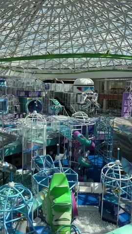 This video shows a large indoor playground with a futuristic theme, featuring numerous interconnected structures, slides, and ball pits. The playground is predominantly colored in shades of blue, purple, and white, with accents of green and silver. At the top of the structure, there is a white, domed object with robotic features, resembling a helmet or a character's head. The entire playground is enclosed by a geodesic dome-like roof with a visible grid structure. The lighting appears to be natural, coming from the roof, and the overall impression is one of a vibrant and expansive entertainment space designed for children. There are no people visible in the frame.