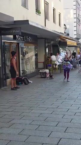 A street performer, dressed in a black tank top, a voluminous purple tutu, and striped leg warmers, is making large bubbles in a paved outdoor walkway. To the performer's left, a second performer, dressed in a black hat, red tank top, and black shorts, stands facing the first performer, holding a long wand. A large black duffel bag and a smaller black bag are on the ground near the second performer. In the background, several people are seated at tables outside a shop with a sign reading "Marc O'Polo." The shop's interior is visible through a large glass window. Buildings line the walkway, with awnings over the shop entrances.