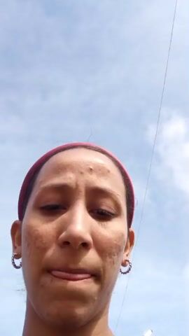 A person is taking a selfie outdoors. They are wearing a black tank top and a pink headband. They have a tattoo on their arm. The background shows a sidewalk, a fence, trees, and a street. The sky is blue with some clouds. The person is puckering their lips and winking at the camera.