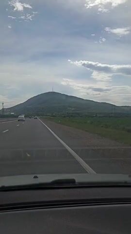 The video is captured from the perspective of someone inside a vehicle, looking forward through the windshield. The road ahead is paved with white lane markings. To the right of the road, there is a grassy field, and beyond that, rolling hills and mountains. Some distant structures, possibly industrial, are visible near the base of the mountains. Power lines and poles are also scattered across the landscape. The sky is partly cloudy with a mix of blue sky and white clouds. The video appears to be taken during daylight hours, possibly late afternoon. There are other vehicles on the road, indicating it is a public highway. The overall scene suggests a journey through a rural or semi-rural mountainous region.