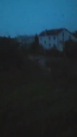 The video opens with a dark, blurry view of what appears to be houses and trees against a deep blue sky. As the video progresses, the sky lightens to a pale gray and the scene becomes clearer, revealing more details of the houses, greenery, and what appear to be raindrops on the camera lens or window. There are no people or clear actions depicted in the video. The environment appears to be a residential area with multiple buildings and foliage, possibly experiencing some form of inclement weather like rain or a storm.