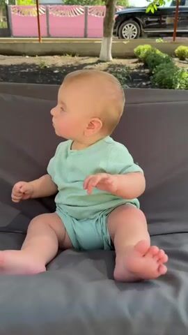 A baby in a light green onesie is on a gray cushioned outdoor swing. The baby is shown sitting up, then turning to look to the side, then reaching out an arm, and finally turning over to lie on its stomach. The baby's legs are visible throughout. The background shows a garden with plants, a pink fence, and a car. Sunlight is casting shadows on the ground.