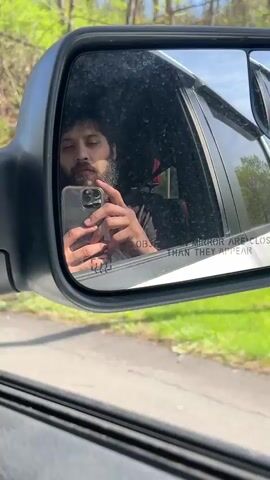 A close-up video shows a man with dark, curly hair and a beard reflected in the driver's side mirror of a vehicle. He is holding a grey smartphone with three cameras in his left hand, appearing to record himself. Initially, he looks directly into the mirror with a neutral expression. He then flexes his right bicep, revealing a tattoo that appears to read "013" on his inner arm. The reflection also shows the partial interior of the car, including a glimpse of a red seatbelt. Outside the vehicle, the background reflects green foliage and trees, indicating an outdoor setting, possibly along a road in Milton, United States of America. The time of day appears to be daytime, with bright natural light. The well-known warning "OBJECTS IN MIRROR ARE CLOSER THAN THEY APPEAR" is visible in white text on the lower right portion of the mirror's surface. The mirror itself has some specks or dust on its surface. The man's expression shifts to one of confidence as he flexes his arm.