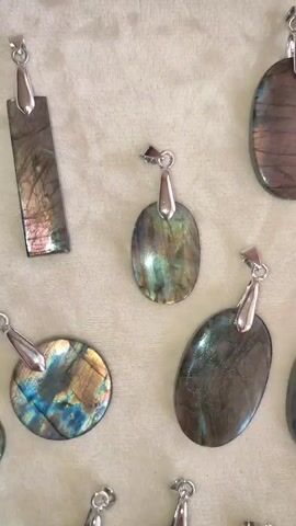 The media file is a close-up, overhead view of a collection of labradorite pendants laid out on a textured, light-colored surface, possibly fabric or a similar material. The pendants are of various shapes and sizes, including oval, rectangular, and round, and each is attached to a silver-toned bail. The labradorite stones exhibit the characteristic iridescence of the mineral, displaying flashes of blue, green, yellow, and purple light as the angle of view changes. Some stones are more translucent than others, revealing internal patterns and textures. The lighting appears to be artificial, highlighting the reflective qualities of the stones. There is no visible text in the media file. The context provided indicates the location is Kriukivshchyna, Ukraine. The scene suggests a display of jewelry for sale or a personal collection.