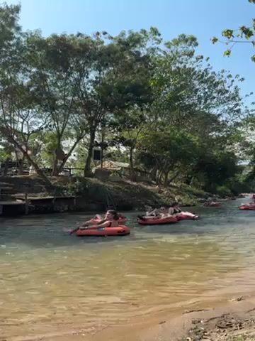 The video shows a slow-moving river with people floating down it in red inner tubes.  The river is surrounded by trees and foliage, and the sky is blue. There is a small building with a dark roof visible on the bank in the distance. The river appears shallow and the water is clear. The people in the tubes are spaced apart and appear to be relaxing.
