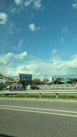 This video shows a view from inside a car, looking out through the windshield. The car is on a road, possibly a highway, with multiple lanes. There is a pedestrian overpass visible on the left side of the road. The sky is blue with scattered white clouds. The sun is shining, casting shadows. There are some trees and buildings visible in the distance. The car is moving forward.