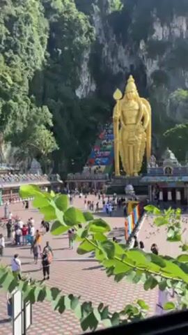 The video shows an outdoor view of the Batu Caves in Malaysia. A giant, golden statue stands prominently in the foreground. Behind the statue, colorful stairs lead up to the cave entrance in the hillside. Many people are walking in the plaza in front of the statue and on the stairs. Green foliage partially obscures the lower left corner of the frame. The weather appears sunny and bright.