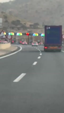 The video shows a highway toll plaza. A large blue truck is in the foreground, driving towards the toll booths. Several cars are in front of the truck, queuing to pay the toll. The toll booths have colored lights indicating their status, with some showing green and others red. The road is paved and has white lane markings. In the background, there are hills or mountains, and the sky appears overcast. The overall scene is that of a typical traffic situation at a toll collection point on a highway.
