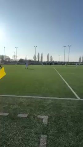The video shows a soccer game from the perspective of the sideline. Players in white uniforms are scattered across a green field under a bright blue sky. There are tall light poles lining the field. A yellow corner flag is visible in the lower left. Cheers erupt after a player scores a goal.