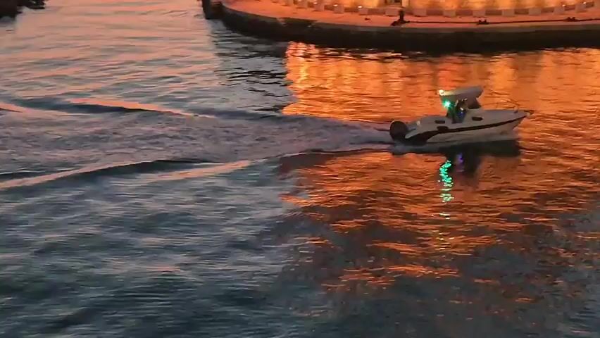 This media file captures a small, white motorboat with a canopy and green lights, cruising on dark blue water. The boat is creating a wake as it moves, and its reflection, along with the reflections of green lights, shimmer on the water's surface. In the background, a structure with warm, orange and yellow lights is visible along a waterfront. The scene appears to be a harbor or bay, possibly at dusk or dawn, given the lighting. The water's surface shows ripples and is influenced by the ambient light, creating a dynamic interplay of colors and textures. The overall mood is tranquil and serene, with the gentle movement of the boat and the soft glow of the lights. There is no visible text in the media file. The location context of Taranto, Italy, suggests this might be a scene along the city's coastline or within its harbor area.
