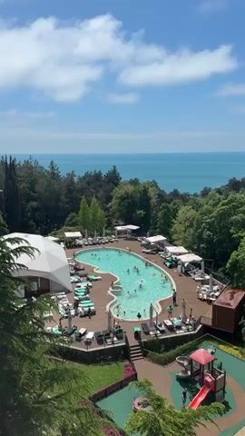 From a high vantage point, the video showcases an outdoor swimming pool surrounded by a wooden deck.  Many people are swimming in the pool, some using inflatable toys.  Around the pool deck, numerous lounge chairs are arranged with some individuals sunbathing or relaxing under umbrellas.  Lush green trees surround the pool area.  In the background, a view of the ocean is visible under a bright blue sky with scattered clouds.  A large white tent-like structure stands on one side of the pool area. People are also walking and standing around the pool area.
