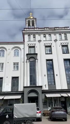The video shows a street scene in front of a white building with a gold cross on top of a bell tower. The building has multiple stories with arched windows on the upper floors and large windows on the lower floors, some of which appear to be storefronts. There are awnings over the storefronts. In front of the building, there are several parked cars and a small truck with a covered bed. The sky is cloudy. The camera is positioned low, looking up at the building. The video is short and shows a slight pan to the right.