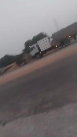 The video, taken from the side of a road, captures a blue tanker truck driving along the road. There are other vehicles and trees visible in the background. The sky is overcast, suggesting it might be early morning or late evening. The roadside is uneven and appears to be partially paved with concrete. There is also a parked car to the left.