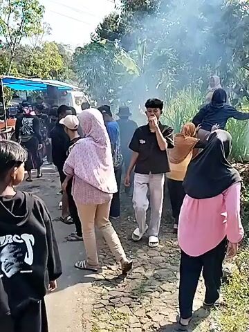 The video depicts an outdoor gathering of people near a road with trees and vegetation. A blue tarp covers a structure that appears to be a makeshift stage or stall. Several individuals are visible, some wearing headscarves and loose clothing. Smoke is present in the air, suggesting a fire or a performance involving smoke. In the latter part of the video, some individuals are engaged in what appears to be a performance or dance, with one person in the foreground wearing traditional attire. The overall atmosphere seems to be one of a community event or celebration.