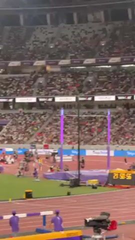 The video shows a male athlete successfully performing a pole vault in a stadium full of spectators. He vaults over the bar without knocking it down, landing safely on the cushioned mat. The stadium is packed with people filling the stands, and a digital clock displays the time: 21:51:01. Several cameras and personnel are visible around the track area.