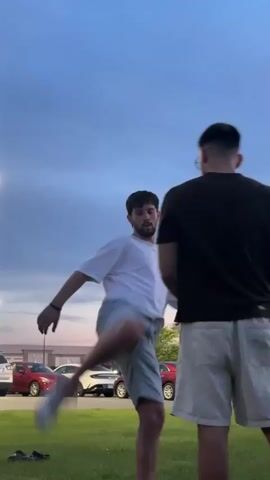 The video shows two men standing in a grassy area with cars parked in the background. The man on the left, wearing a white t-shirt and denim shorts, performs a martial arts-like kick and then a series of hand movements. The man on the right, wearing a black t-shirt and white shorts, stands facing him, seemingly observing. The sky is a dusky blue with some clouds. The lighting suggests it is either dusk or dawn.
