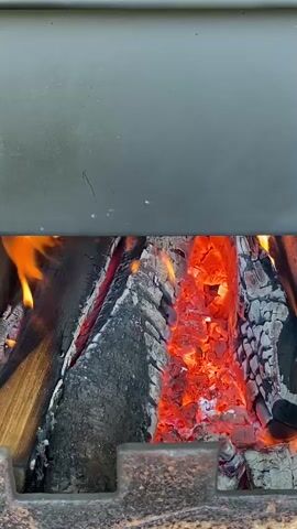 A close-up, stationary shot focuses on the interior of a dark metal firebox where burning wood is actively producing flames and glowing embers. The logs are partially consumed, with charred bark and ash accumulating. Intense orange and yellow flames flicker and dance among the wood, while the underlying coals radiate a deep red glow. The firebox appears to be part of a larger structure, possibly a grill or fireplace, indicated by the dark metal casing above and a textured, dark metal grate or base below. The lighting is natural and primarily sourced from the fire itself, casting a warm, dynamic light on the scene. No people are visible, and the focus remains solely on the burning wood and its immediate surroundings.