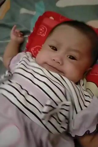 A baby is lying on its back, resting on a red pillow with cartoon faces on it. The baby is wearing a striped onesie and a light purple romper. The background is a bed with a patterned sheet. The baby's head is tilted slightly to the left, and its eyes are open and looking towards the camera. The baby's mouth is slightly open, and it appears to be making a sound. The baby's left hand is extended upwards and slightly to the left. The video is a close-up shot of the baby.
