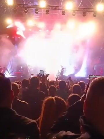 The video shows a large crowd of people at a concert, from a lower perspective in the audience. Many are holding up phones to record the show. A dense cloud of white confetti is released into the air over the crowd, illuminated by bright stage lights in red, white, and blue. The stage setup includes patterned light panels. People are cheering and appear excited.