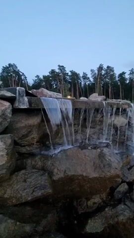 A man and a woman in white robes stand on a wooden walkway next to a small man-made waterfall. The waterfall cascades over rocks into a pool of water. Pine trees line the background of the scene. The sky is a pale blue, indicating it is either dawn or dusk.