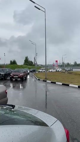 The video shows a gas station with several cars in line. The weather appears to be overcast and rainy, as evidenced by the wet pavement and water droplets on the cars. The cars are of various makes and models, including SUVs and sedans. Some people can be seen inside the vehicles. The gas station has red and white branding with text in Russian. The scene suggests a normal operation of a gas station, possibly with moderate traffic.