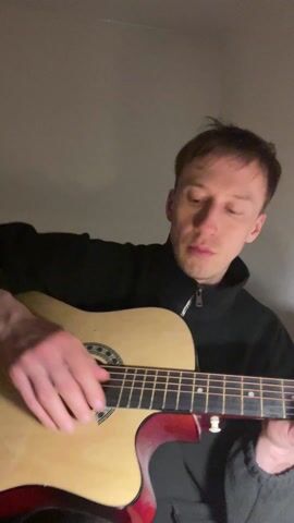 The video shows a man playing an acoustic guitar. He is indoors, against a plain light gray wall, wearing a black zip-up fleece jacket. He looks directly at the camera while holding the guitar, and the middle finger of his left hand is extended upward while he strums the strings with his right hand. The guitar is light-colored with a red back and sides, and the focus is on the man's face and hands. The lighting is warm, and the overall impression is intimate and confrontational.