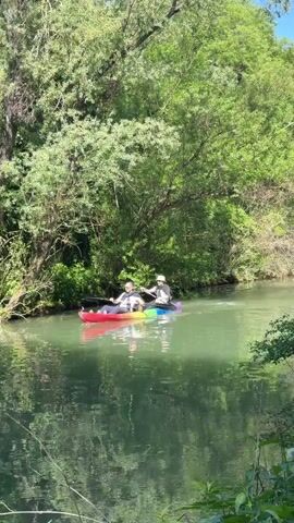 The video shows a tranquil river scene with lush green trees lining the banks. A rainbow-colored kayak with two people paddling gently glides through the water. Nearby, a white paddleboat with four individuals, some relaxing, adds to the serene atmosphere. The calm, slightly murky water reflects the surrounding greenery, creating a peaceful and picturesque scene.
