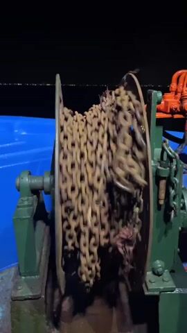 This video shows a close-up view of a large, rusty chain being wound onto a winch. The winch is green and appears to be made of metal. The chain is heavy and appears to be made of thick links. The background of the video is dark, and it is difficult to see what is in the background. The winch is on a boat or ship. The chain is moving quickly.
