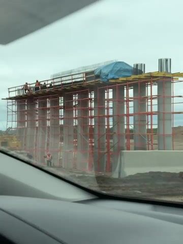The video shows a view from inside a vehicle looking out at a construction site. A large structure, likely a bridge support, is under construction with scaffolding and concrete pillars. A "no entry" sign and a detour sign are visible. Piles of sand and gravel are present, indicating ongoing earthwork. Several other vehicles, including a silver SUV, a dark sedan, and a blue hatchback, are also visible on the road. Power lines and utility poles are scattered throughout the landscape, which appears to be a somewhat barren and industrial area under a cloudy sky.