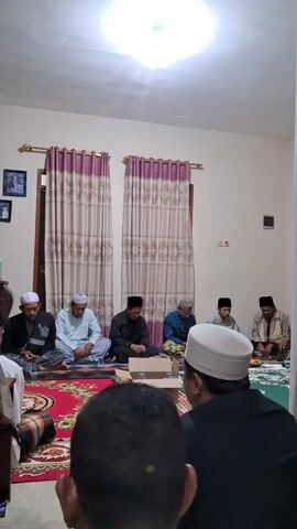In a medium shot under a bright, diffused light, seven men are seated on the floor in a carpeted room, with their focus directed towards an unseen speaker. The men are attired in a variety of modest clothing, with several wearing traditional Muslim head coverings, including white caps. Some are sitting on patterned rugs, and there is a small cardboard box and a plastic water bottle visible in the center of the gathering. Behind them, a window is flanked by two patterned curtains in shades of pink and beige. Photographs hang on the wall to the left of the window. The room appears to be a simple living space, and the atmosphere seems to be one of respectful attention to whoever is speaking.
