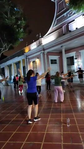 At night, a group of people are exercising in front of a light pink building with columns. The building has a sign that says “BIBLIO” above the entryway. A tree is to the left, and there are several water bottles on the ground. The floor is covered in a red tile pattern. The group of people are moving their arms in a coordinated fashion. There are approximately ten people in the group, and they are wearing a variety of casual clothing. The scene is well-lit, and the overall atmosphere appears to be positive and energetic.