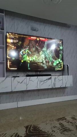A flat-screen TV, showing a video game fight scene, is mounted above a white marble-patterned media console against a vertically striped wall. There is a bookshelf with decorative items on the left side of the TV, and a Playstation 5 console on the right.  The floor is shiny with a marbled pattern. The scene is bright, and a round light fixture is visible on the ceiling.