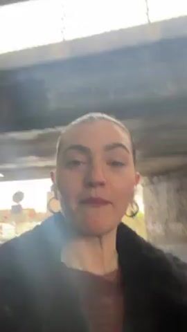 The video shows a woman walking underneath an overpass and talking to the camera. She is wearing a dark brown top and a black jacket. She has large hoop earrings. Her hair is pulled back. The woman says she is pro on "yapping," if that counts. The video zooms in closely on her face. Graffiti can be seen on a wall in the background. The camera view is shaky.