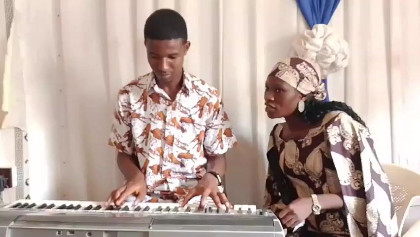 Two individuals are featured in the video: a young man playing a keyboard and a woman standing next to him, presumably singing. The man is wearing a patterned short-sleeved shirt and a watch. The woman is wearing a patterned dress with a matching headscarf. She also has a watch on.

The man is focused on playing the keyboard. The woman is looking towards the camera, seemingly singing along with the music.

They are positioned in front of a white curtain backdrop, decorated with a blue drape and a white floral arrangement. A wall outlet is visible to the left. A white plastic chair is on the right. The setting appears to be indoors, potentially in a church or performance space.