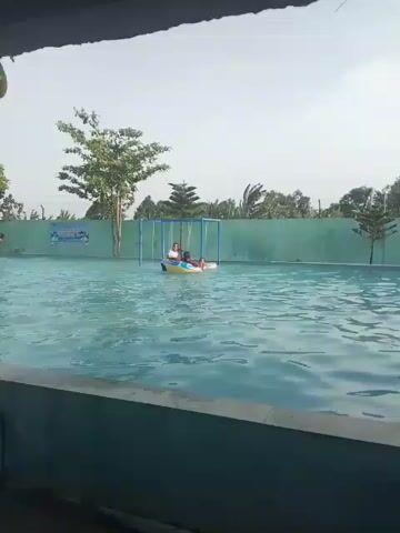 The video shows an outdoor swimming pool on a cloudy day. The pool is light blue, with ripples on the surface. In the middle of the pool, a few children are in inflatable tubes. One tube has three children in it, while another has one child swimming next to it. There are several trees and a green wall surrounding the pool area. The camera appears to be positioned at the edge of the pool, looking out over the water.