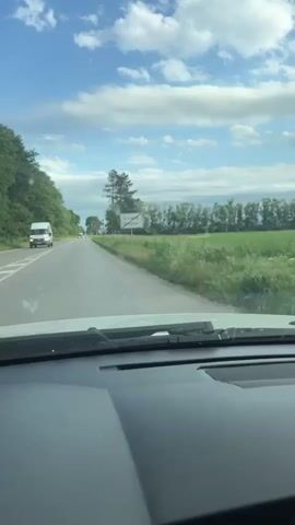 A person drives on a rural road. A sign, partially obscured, is on the right side of the road. Trees border the left side of the road, and a field is visible on the right. The sky is blue with some clouds. The view is from the driver’s seat, looking out the windshield. The person is recording from the rearview mirror, where they can be seen holding up their iPhone. The phone is affixed to a mount attached to the mirror. Other cars are visible on the road ahead.