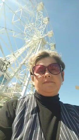 A woman with short gray hair, wearing large pink sunglasses, a black turtleneck, and a pinstriped vest, takes a selfie in front of a large white Ferris wheel on a sunny day. The Ferris wheel is the most prominent object in the background, with several gondolas visible. To the left and right of the Ferris wheel, there are glimpses of amusement park rides and structures, including a green and white carousel-like ride and a red and white building with an arched entrance. People are visible in the background, walking around the park. The sky is clear and blue.