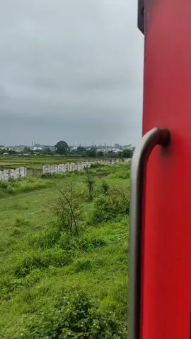 The video shows a view from a moving train. The train car is red and a metal handle is visible on the right side of the frame. Outside the train, there is lush green vegetation, trees, and a cloudy sky. In the distance, some buildings and structures can be seen. The train appears to be moving along a track next to a grassy area and a short fence. There are no people visible in the frame.