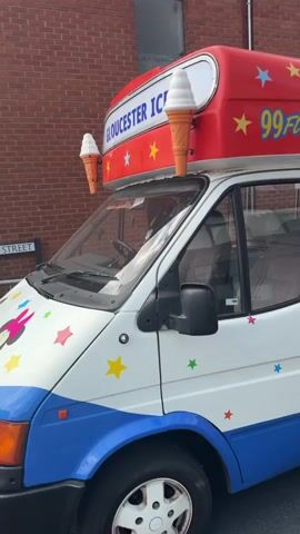 The video shows an ice cream truck with "Gloucester Ices" and "99 Flake" written on its side. The truck is parked on a street, which appears to be closed off for an event. There are colorful flags strung across the street, and several tents and food stalls are visible in the background. A few people are walking around, and there are trash bins and what looks like a beer keg near one of the tents. The scene suggests a street fair or a local event.