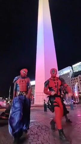 At night, two people dressed in Spider-Man and Deadpool costumes are dancing in front of a tall white obelisk lit with purple and pink lights. A third person in a superhero costume joins them. They are on a stone plaza in a city. There are some cars, streetlights, and a building with an advertisement in the background. The sky is dark.