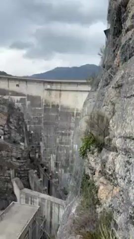 The video shows a very high concrete dam in a mountainous area. The camera is positioned high up, offering a downward view of the dam and the surrounding landscape. The dam is massive and appears to be built in a steep canyon. Below the dam, there are concrete structures, likely associated with the dam's operations, including a building and what seems to be a reservoir or a settling basin with greenish water. The sky is overcast, suggesting a cloudy day. There is a system of wires and cables in the foreground, possibly part of the infrastructure related to the dam. A group of people can be seen swimming in the pool.