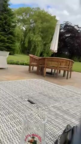 Here is a description of the video:

The video starts with a close-up shot of a woven outdoor table with a glass on it. The perspective is low, looking through what appears to be someone's legs. The camera angle shifts to show the person's legs extended in front of them, revealing they are wearing light-colored pants and blue socks with black stripes and red toes. The socks feature images of London buses. The background shows an outdoor patio with wooden benches, a white umbrella, green trees, and a cloudy sky. The camera remains steady, focusing on the person's legs and the view of the yard.
